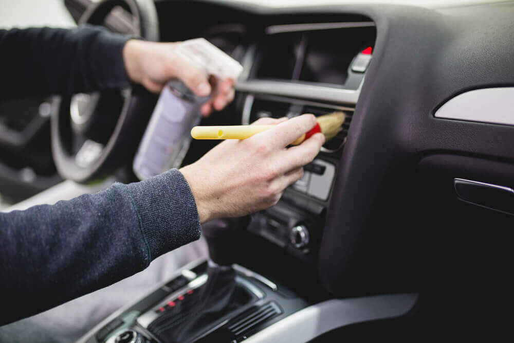 an interior detail cleaner cleaning the interior console of a car with a dust removal brush and holding a spray bottle