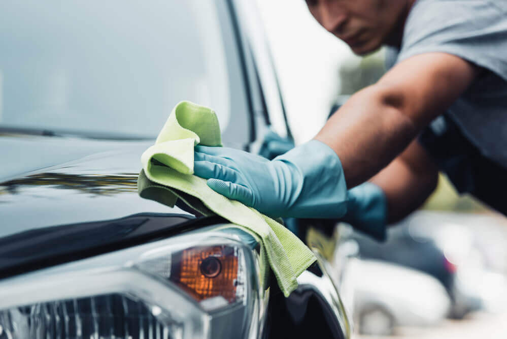 an exterior detail cleaner wearing protective gloves wiping down the front panel of a black car with a clean cloth 
